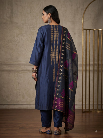 A woman stands in an ornate stone archway wearing a traditional Indian outfit consisting of a navy blue kurta with gold zari embroidery, matching navy blue pants, and a sheer navy dupatta with purple and gold borders.