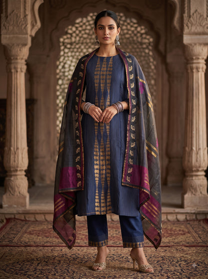 A woman stands in an ornate stone archway wearing a traditional Indian outfit consisting of a navy blue kurta with gold zari embroidery, matching navy blue pants, and a sheer navy dupatta with purple and gold borders.