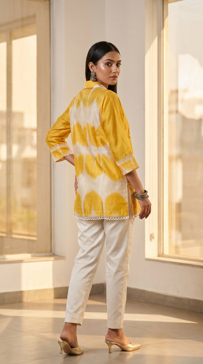 Woman wearing a yellow top and white pants in a bright room with large windows.